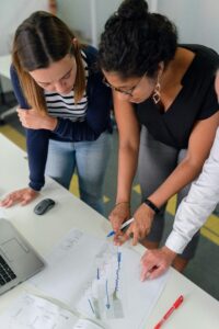 two students leaning over a table in front of a computer writing on white paper